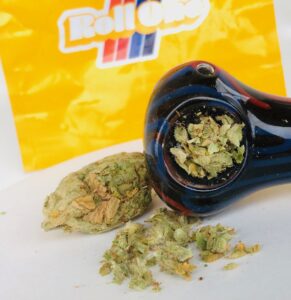 glass pipe tilted on its side revealing a bowl ground lemon kush headband with bud and other ground bits of lemon kush headband in front of a rollone ziplock bag